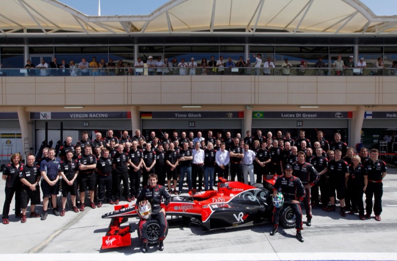 Virgin Racing team members in black and red uniforms gathered around their car and two drivers in the Bahrain pit lane.